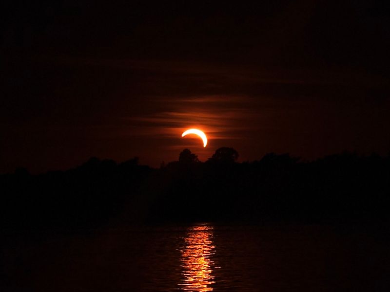 Częściowe zaćmienie Słońca nad Jeziorem Dominickim: sierp Słońca nad linią lasu i jego odbicie w wodzie